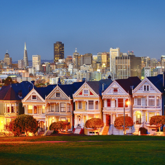 The Painted Ladies of San Francisco, California sit glowing amid the backdrop of a sunset and skyscrapers.
