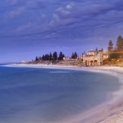 Perth western Indian ocean facing sandy beach Cottesloe at sunset. Central pavilion with lights and trees above blurred waves and water.