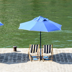 Three blue parasols with blue and white striped deck chairs in the sun on the wharf of the river Seine with green water.