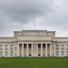 Auckland, New Zealand - March 1, 2017: The beige, completely deserted north facade of the rectangular War Memorial Museum between the green lawn and a dark gray cloudy sky. Lanterns hold burning lamps