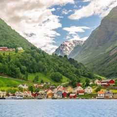 A lots of little colorful traditional Norwegian houses near the sea with green forest on mountains on background and blue sky with dark clouds, Sognefjord, Norway