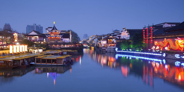 图片展示了夜幕下的中国古城河流两岸，亮起了色彩缤纷的灯光，有船只停泊，古典建筑映衬着现代城市轮廓。
