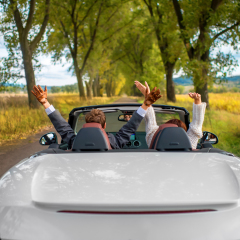 smiling couple sitting in their white car on the road with hands uo