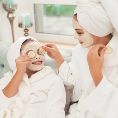 Photo of mother and daughter in white bathrobes. They are sitting on the couch and applying pieces of cucumber to their eyes. Their hair is wrapped in a white bath towel. They have cucumber mask.