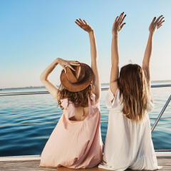 Outdoor back shot of two young female on luxury vacation, waving at seaside while sitting on yacht. Best friends are waiting for waiter to order some drinks as they are sailing on cruise ship