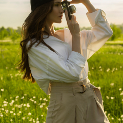 Beautiful girl photographer wearing a white shirt, beige trousers poses outdoors with a vintage camera against a background of meadow full of flowers in sunset light. Lifestyle design. Copy space.